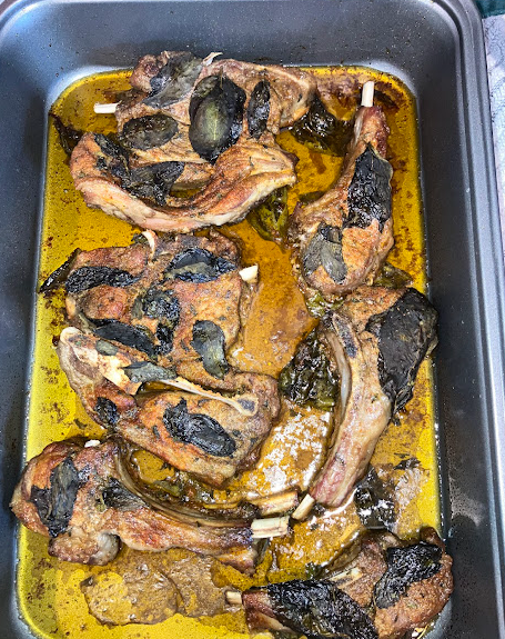 Cooked lamb chops and shoulder steaks in a pan with fresh mint leaves spread around the top and olive oil and seasonings on the pan